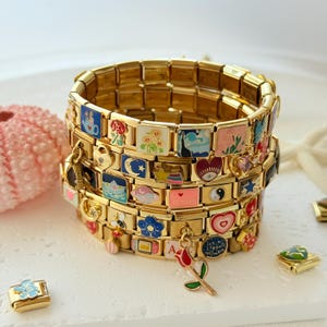 May include: Gold-toned charm bracelets stacked on a white surface. Each bracelet link features a colorful enamel charm with various designs, including flowers, hearts, and celestial motifs. A rose charm hangs from one bracelet. Additional charms are scattered around.
