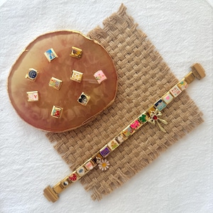 May include: A gold charm bracelet with a variety of colorful charms, including flowers, hearts, and a four-leaf clover. The charms are arranged in a row on the bracelet, which is lying on a brown woven surface. There is a separate set of charms on a pink and gold surface.