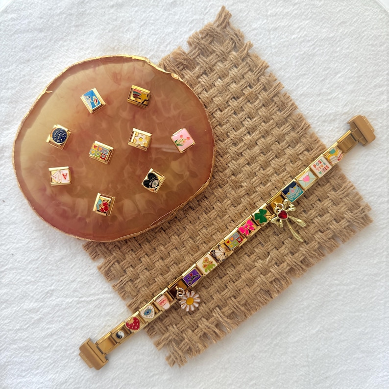 May include: A gold charm bracelet with a variety of colorful charms, including flowers, hearts, and a four-leaf clover. The charms are arranged in a row on the bracelet, which is lying on a brown woven surface. There is a separate set of charms on a pink and gold surface.
