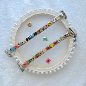 May include: Two silver charm bracelets with colorful enamel charms featuring various designs like hearts, flowers, and smiley faces. The bracelets are displayed on a white surface with a decorative hoop and scattered charms.