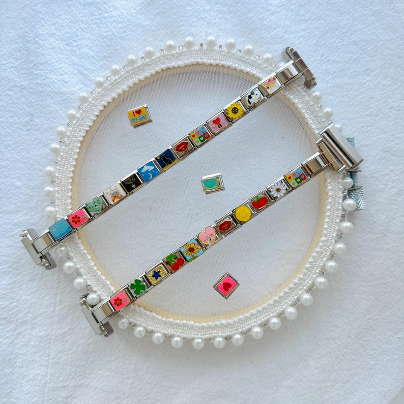 Pu&ograve; includere: Due braccialetti con ciondoli in argento con ciondoli smaltati colorati con vari disegni come cuori, fiori e faccine sorridenti. I braccialetti sono esposti su una superficie bianca con un cerchio decorativo e ciondoli sparsi.