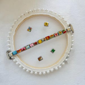Op de afbeelding: Een zilverkleurige modulaire schakelarmband met kleurrijke emaille bedels, waaronder een zonnebloem, kersen, een hartje, een regenboog en een smiley, geplaatst op een ronde witte borduurring versierd met een rand van kleine witte parels.