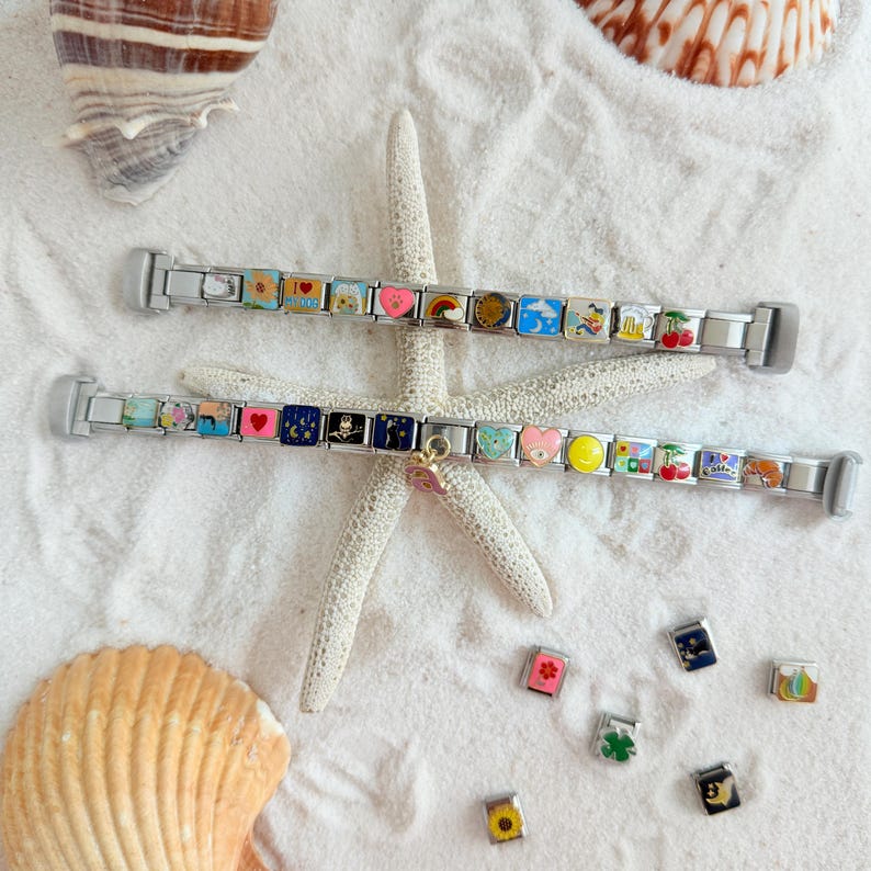 Pu&ograve; includere: Due braccialetti con ciondoli argentati con ciondoli smaltati colorati raffiguranti cuori, arcobaleni e altri disegni. I braccialetti sono esposti su sabbia bianca con una stella marina e conchiglie.