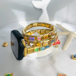 Op de afbeelding: Goudkleurige bedelarmbanden met kleurrijke emaille bedels, waaronder harten, koffiekopjes en een zonnebloem. Een zwarte smartwatch met een gouden band is bevestigd aan een armband. De armbanden zijn op een wit oppervlak gestapeld.
