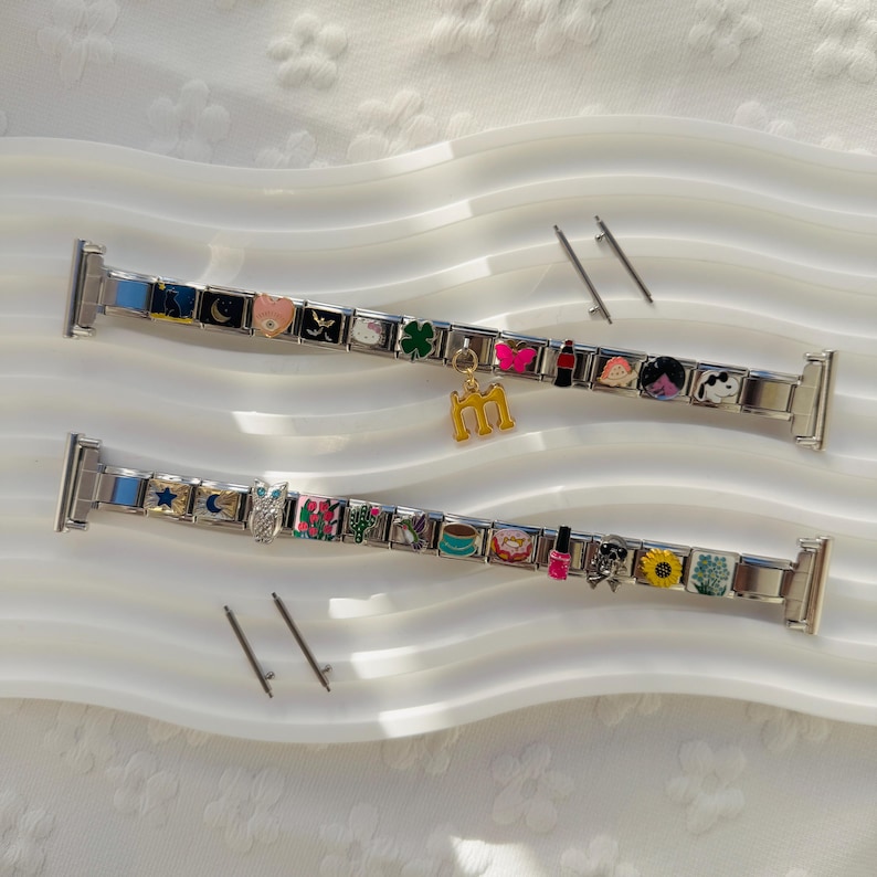 May include: Two silver-toned modular watch bands featuring a series of colorful enamel charms including a moon, clover, butterfly, coffee cup, donut, and sunflower. One band includes a gold-toned letter M charm hanging from a central link.