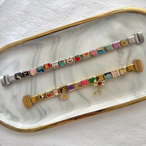 May include: Two charm bracelets with colorful enamel charms. The top bracelet is silver with a sunflower charm and the bottom bracelet is gold with a daisy charm.