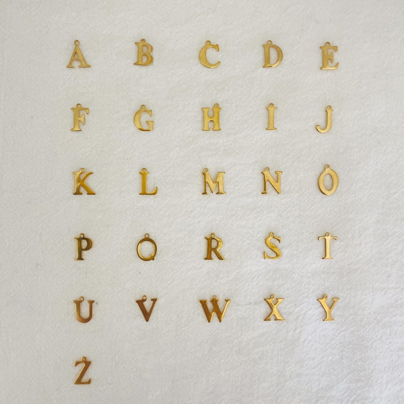 May include: A selection of gold-coloured alphabet charms, each with a small loop at the top for attaching to a chain or bracelet. The letters are displayed on a white, textured surface.