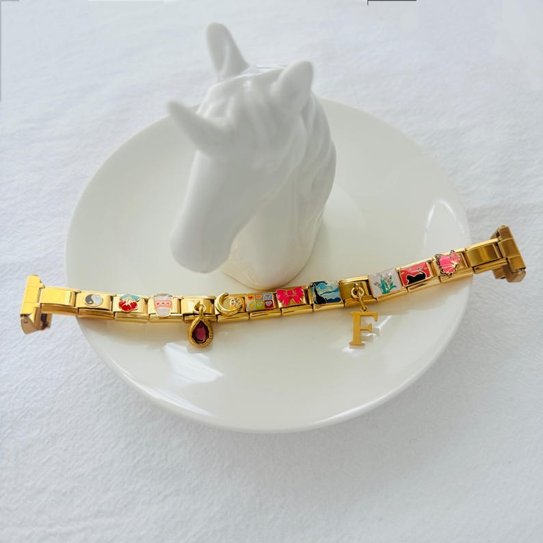 May include: A gold charm bracelet with colorful charms, including a yin yang symbol, a strawberry, a cup of coffee, a crescent moon, a flower, a cat, and a letter "F".