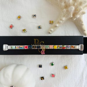Pu&ograve; includere: Un braccialetto con ciondoli in argento con ciondoli in smalto colorato, tra cui un cuore, un orso e una ciliegia. Un ciondolo rosa con la lettera "a" pende dal braccialetto. Diversi ciondoli sciolti sono sparsi.