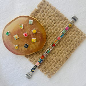 Puede incluir: Un brazalete de plata con abalorios de esmalte de colores que presentan varios diseños, incluyendo flores, estrellas y formas geométricas. Los abalorios se muestran sobre un fondo beige.