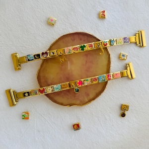 May include: Two gold charm bracelets with colourful enamel charms, including hearts, stars, and flowers. The bracelets are displayed on a decorative surface with scattered charms. The charms have various designs and the bracelets have a clasp closure.