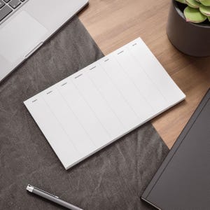 May include: A white weekly planner pad with vertical columns for each day of the week, labeled Sunday through Saturday, plus a notes section. The pad is on a wooden desk with a laptop, pen, succulent plant, and a black notebook.