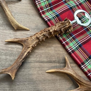 May include: A bottle opener with a polished silver metal opener and a brown antler handle. The antler handle is textured and has a natural finish. The bottle opener is lying on a red and green tartan fabric.