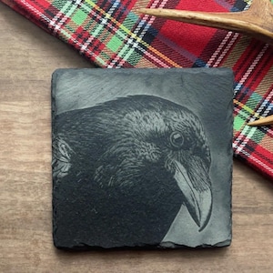 Op de afbeelding: Een zwarte leisteen onderzetter met een gedetailleerde gravure van een ravenkop. Het oog van de raaf is zichtbaar en zijn snavel is lichtjes open.
