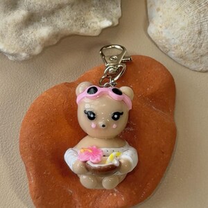 Könnte beinhalten: Ein niedlicher Bären-Schlüsselanhänger mit rosa Visier, der eine Schüssel mit einer Blume und einem Getränk hält. Der Bär trägt ein weißes Outfit. Der Schlüsselanhänger liegt auf einem orangefarbenen Stein, mit Muscheln im Hintergrund.