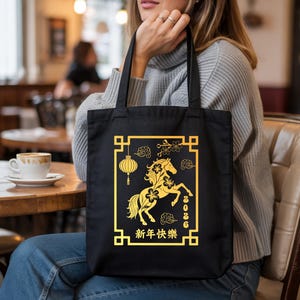 May include: Black tote bag featuring a gold design for the Chinese New Year. The design includes a horse, lantern, and Chinese characters. The year 2026 is also displayed. The bag is held by a person.