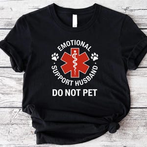 May include: Black t-shirt with the text "EMOTIONAL SUPPORT HUSBAND" surrounding a red medical symbol and paw prints. Below the text is "DO NOT PET". The shirt is made of a soft material.