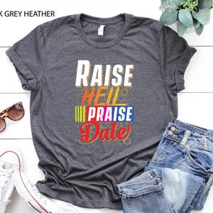 May include: Dark grey heather t-shirt with the text "Raise Hell Praise Dale" in colorful letters.