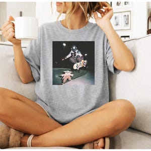 May include: A heather grey short-sleeved t-shirt featuring a square graphic print on the chest. The graphic depicts a figure in a dark helmet and armour performing a skateboard trick in the air, while a small green creature stands on a skateboard.