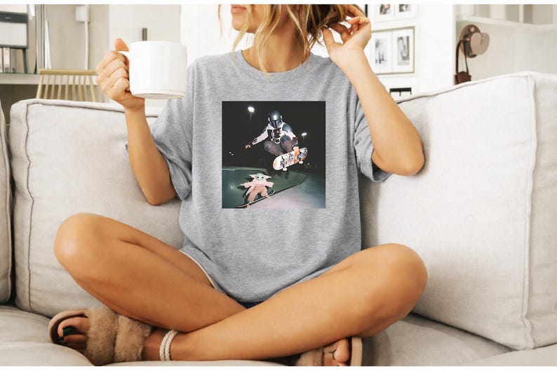 May include: A heather grey short-sleeved t-shirt featuring a square graphic print on the chest. The graphic depicts a figure in a dark helmet and armour performing a skateboard trick in the air, while a small green creature stands on a skateboard.