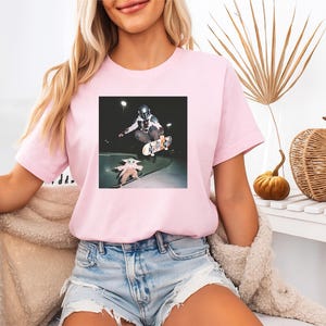 May include: A light pink short-sleeved t-shirt featuring a graphic print of a character in a helmet riding a skateboard and a small creature on a separate skateboard. The design is centred on the chest of the shirt, which has a crew neckline.