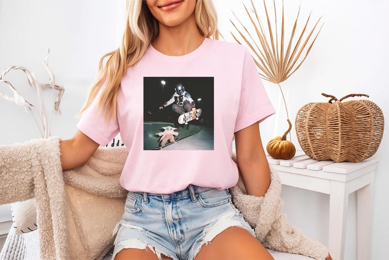 May include: A light pink short-sleeved t-shirt featuring a graphic print of a character in a helmet riding a skateboard and a small creature on a separate skateboard. The design is centred on the chest of the shirt, which has a crew neckline.