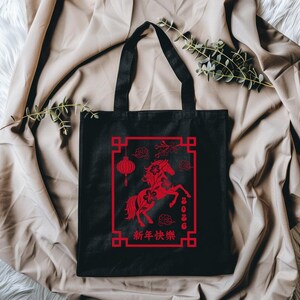 May include: Black tote bag featuring a red graphic of a horse, lantern, and floral elements within a rectangular frame. The text "2026" and Chinese characters are also visible. The bag has black handles and is made of a sturdy fabric.