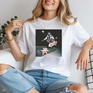 May include: A white t-shirt featuring a graphic print of a character in a silver helmet and armour performing a skateboard trick in the air, while a small green creature in a beige robe stands on a skateboard on the ground below at night.