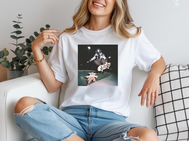 May include: A white t-shirt featuring a graphic print of a character in a silver helmet and armour performing a skateboard trick in the air, while a small green creature in a beige robe stands on a skateboard on the ground below at night.