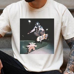 May include: A cream-coloured short-sleeve t-shirt featuring a graphic print of a character in a metallic helmet and armour performing a skateboard trick in a bowl, with a smaller green creature on a skateboard in the foreground at night.