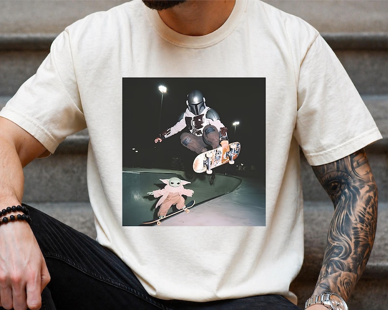 May include: A cream-coloured short-sleeve t-shirt featuring a graphic print of a character in a metallic helmet and armour performing a skateboard trick in a bowl, with a smaller green creature on a skateboard in the foreground at night.