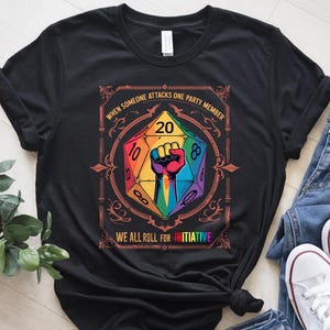 May include: Black t-shirt featuring a colorful D20 die graphic with a raised fist and the text "WHEN SOMEONE ATTACKS ONE PARTY MEMBER WE ALL ROLL FOR INITIATIVE". The graphic is in rainbow colors.