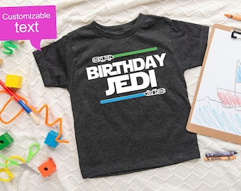 Könnte beinhalten: Ein dunkelgrau meliertes Kurzarm-T-Shirt mit dem weißen Schriftzug "BIRTHDAY JEDI" mittig auf der Brust. Über dem Text befindet sich eine grüne Lichtschwert-Grafik und unter dem Text eine blaue Lichtschwert-Grafik, beide mit detaillierten Griffen.
