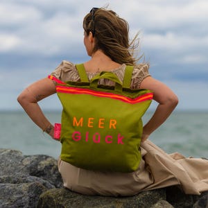 Könnte beinhalten: Eine olivgrüne Tragetasche mit Rucksackträgern, die den Schriftzug MEER GLÜCK in orangefarbenen und pinken Buchstaben trägt. Die Tasche hat einen leuchtend orange-pink gestreiften Rand an der Oberseite und ein kleines pinkes Etikett am Seitengurt.
