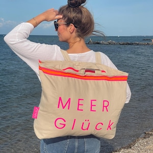 Può includere: Una borsa tote beige con le parole "MEER GLÜCK" in rosa acceso. La borsa ha una striscia arancione e rosa brillante sulla parte superiore. La persona nell'immagine indossa una camicia bianca a maniche lunghe e jeans blu.