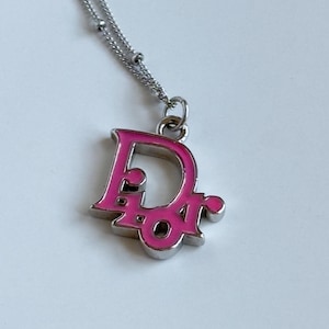 May include: A silver-toned necklace with a pink pendant featuring the letters "Dior". The pendant is filled with pink enamel and hangs from a delicate chain. The necklace is displayed on a white surface.