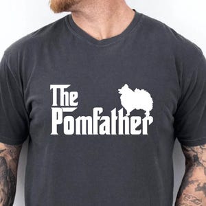 May include: Dark gray t-shirt with the text "The Pomfather" in white, featuring a silhouette of a Pomeranian dog. The shirt has a classic crew neck and short sleeves.