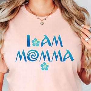 Pode incluir: Uma camiseta rosa com o texto "I am Momma" em azul com um redemoinho azul e verde e duas flores de hibisco azuis.