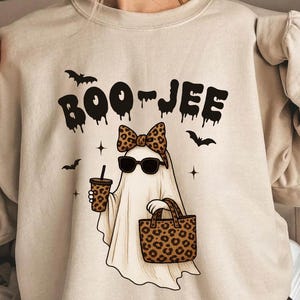 May include: Beige sweatshirt with a Halloween-themed graphic. The design features a ghost wearing sunglasses and a bow, holding a coffee cup and a leopard print bag. The text "BOO-JEE" is above the ghost, with bats and stars.