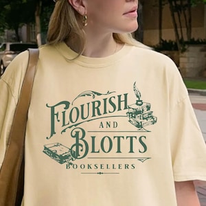 May include: Butter-colored t-shirt with the text "Flourish and Blotts Booksellers" in green. The design includes illustrations of books and a quill. The shirt is part of a casual wear collection.