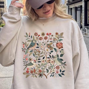 May include: A beige crewneck sweatshirt featuring a colorful botanical illustration of birds and mahjong tiles. The sweatshirt is paired with a beige and grey baseball cap with the text The Western House Casino Las Vegas Nevada printed on the front.