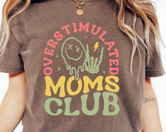 May include: Espresso-colored t-shirt with the text "OVERSTIMULATED MOMS CLUB" in a retro design. The graphic includes a melting smiley face, a skeleton hand, and a lightning bolt. The shirt is paired with blue jeans.