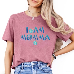 Pode incluir: Uma camiseta rosa com o texto "I am Momma" em azul com um gr&aacute;fico de flor azul de cada lado do texto.