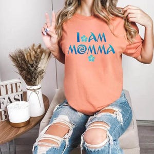 Pode incluir: Uma camiseta cor de p&ecirc;ssego com o texto "I am Momma" em letras azuis e turquesa. O texto inclui uma flor azul e um design de redemoinho turquesa.