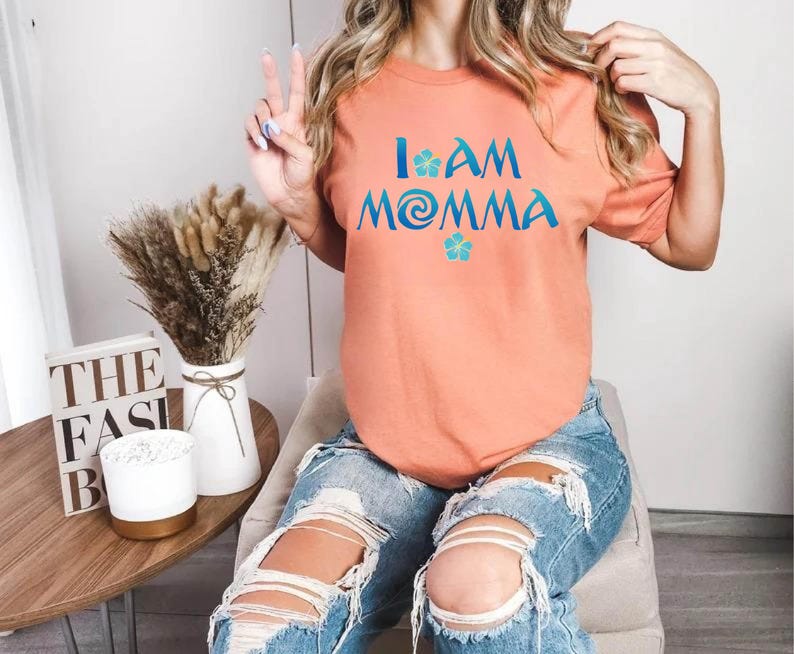 Pode incluir: Uma camiseta cor de p&ecirc;ssego com o texto "I am Momma" em letras azuis e turquesa. O texto inclui uma flor azul e um design de redemoinho turquesa.