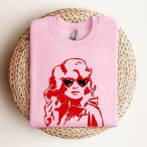 May include: Pink sweatshirt with a red graphic of a woman with blonde hair wearing heart-shaped sunglasses.