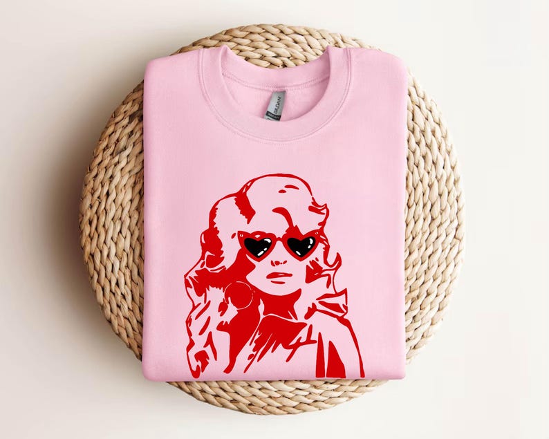 May include: Pink sweatshirt with a red graphic of a woman with blonde hair wearing heart-shaped sunglasses.