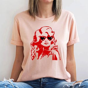 May include: A pink t-shirt with a red silhouette of a woman with large heart-shaped sunglasses. The woman has long, flowing hair.