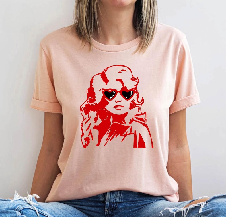 May include: A pink t-shirt with a red silhouette of a woman with large heart-shaped sunglasses. The woman has long, flowing hair.