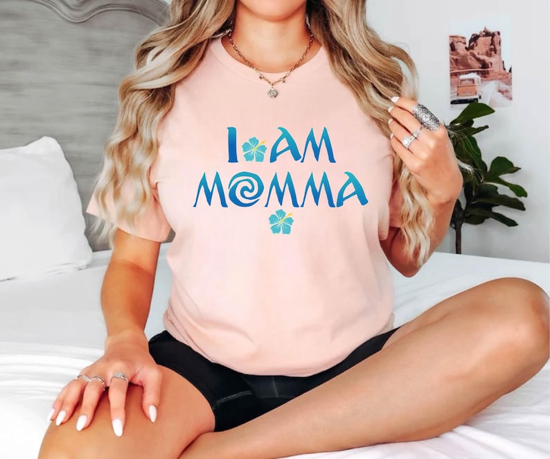Pode incluir: Uma camiseta rosa com o texto "I am Momma" em azul com um redemoinho azul e verde e duas flores de hibisco azuis.